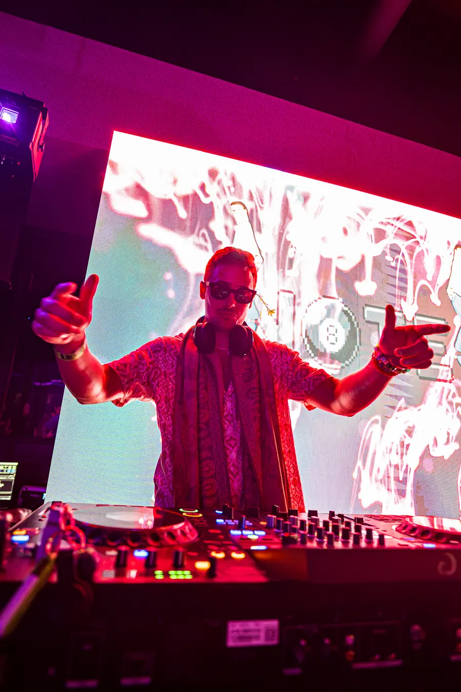 DJ in sunglasses pointing at the crowd from the booth under pink light