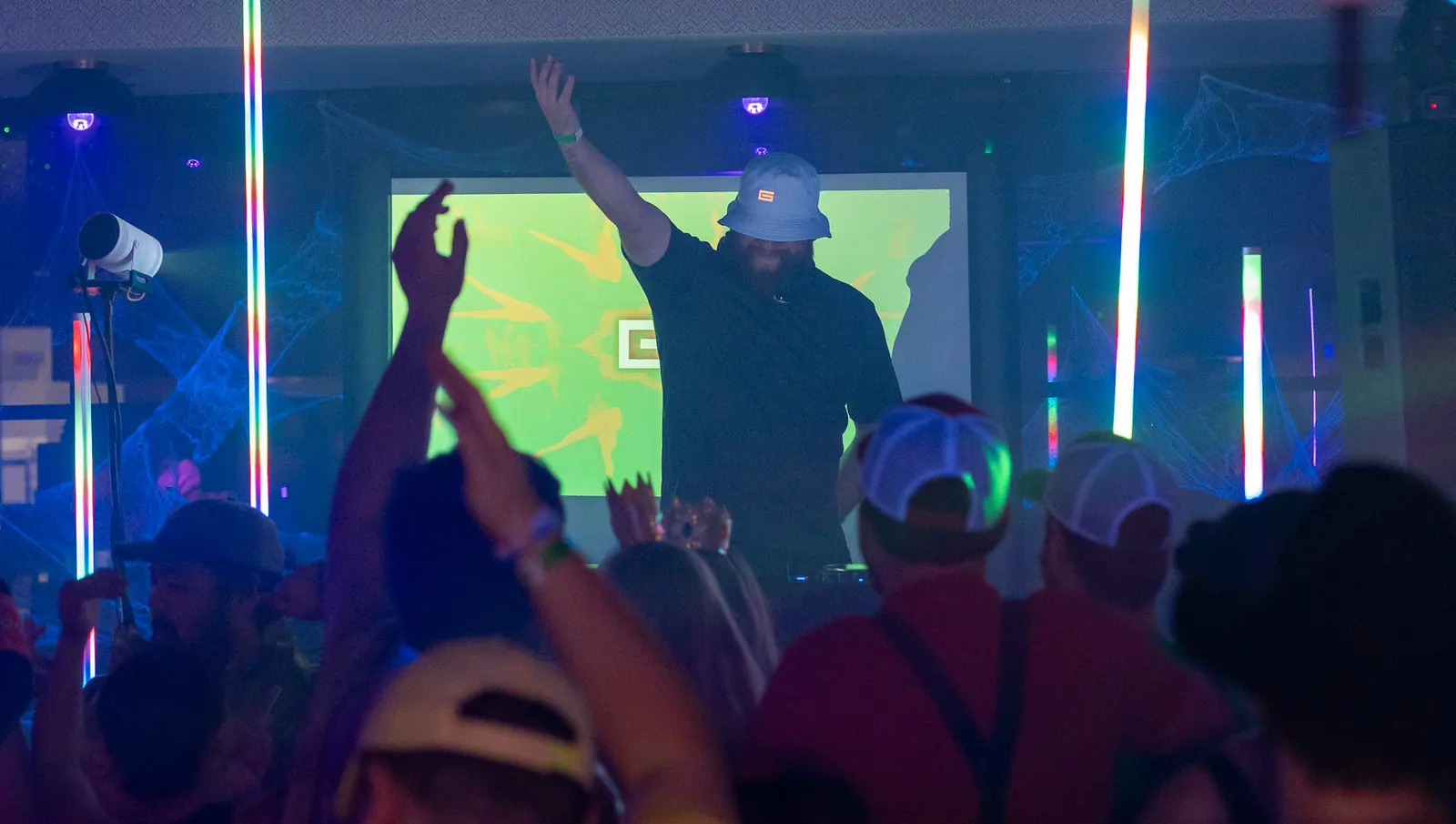 Hands up across a costumed crowd in blue light, red shirts with suspenders, white trucker caps and a crown, as the DJ raises an arm