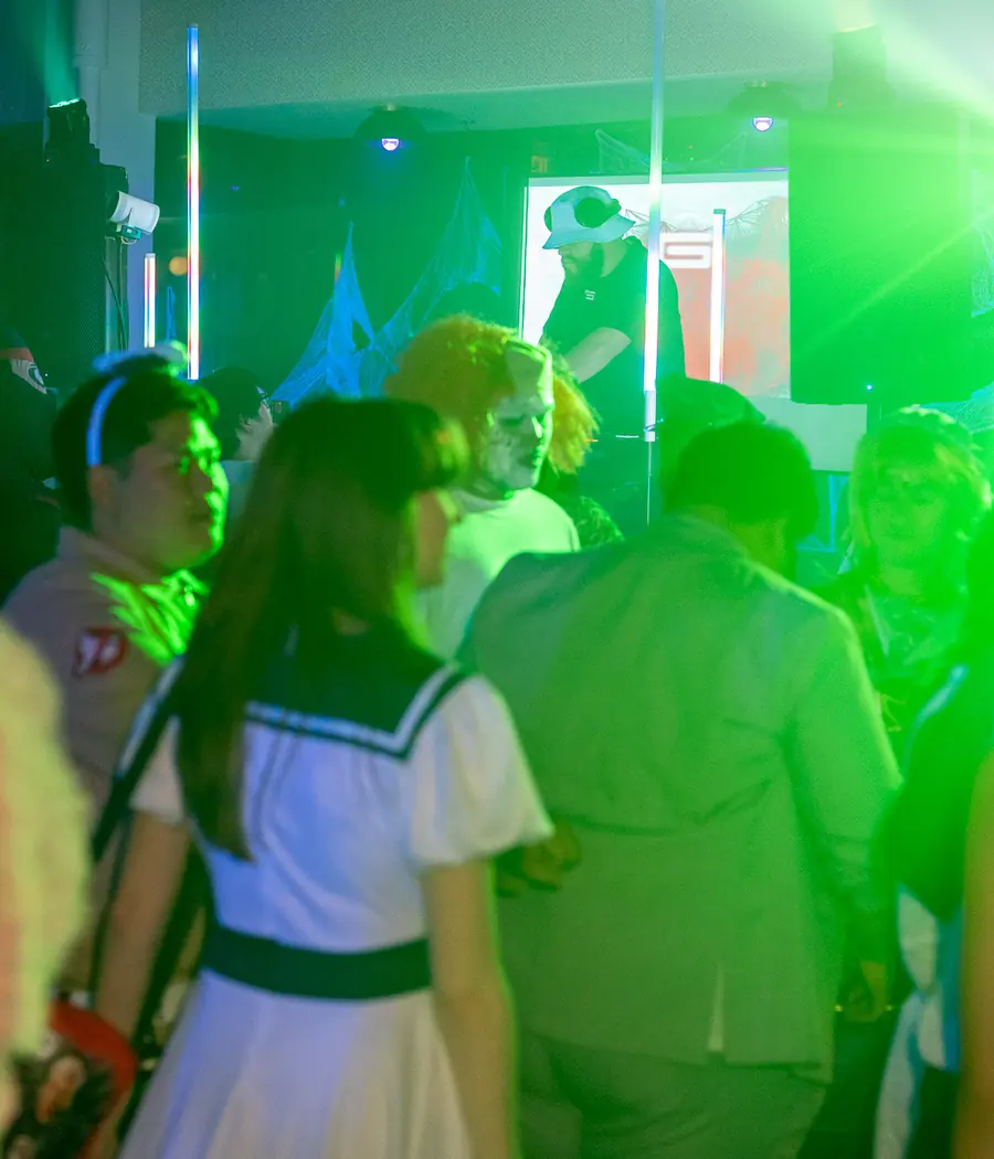 A costumed crowd on the floor in green laser light: a sailor uniform, a ghost-hunter jumpsuit and an orange clown wig with a zombie mask, with the DJ playing behind them